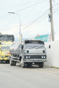 A large gray truck with a tanker attached is parked on a road next to a wall with safety signage. A yellow truck is visible behind it. Overhead, multiple power lines are strung between utility poles.