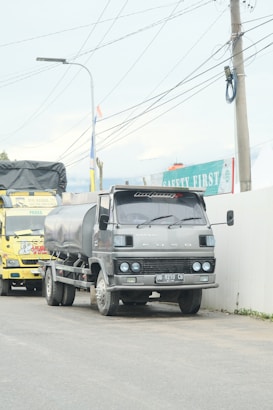 A large gray truck with a tanker attached is parked on a road next to a wall with safety signage. A yellow truck is visible behind it. Overhead, multiple power lines are strung between utility poles.
