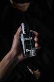 A hand holding a dark, sleek bottle of perfume labeled 'Sauvage' by Dior. The background is dark and creates a moody, sophisticated atmosphere.