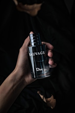 A hand holding a dark, sleek bottle of perfume labeled 'Sauvage' by Dior. The background is dark and creates a moody, sophisticated atmosphere.