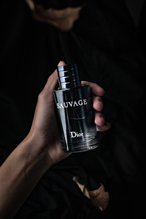 A hand holding a dark, sleek bottle of perfume labeled 'Sauvage' by Dior. The background is dark and creates a moody, sophisticated atmosphere.