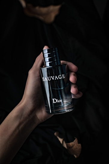 A hand holding a dark, sleek bottle of perfume labeled 'Sauvage' by Dior. The background is dark and creates a moody, sophisticated atmosphere.