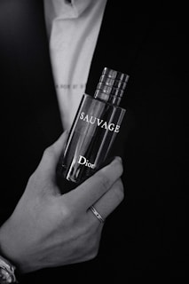 A monochrome image features a hand holding a perfume bottle labeled 'Sauvage' by Dior. The person is dressed in formal attire, including a suit and watch.