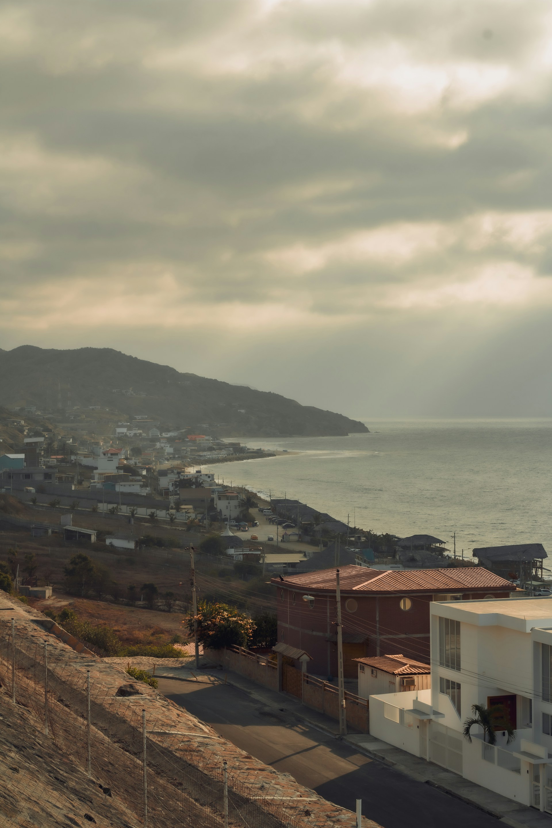 A coastal scene depicting a serene seaside town with houses lining the shoreline. The sky is partly cloudy, and the sunlight gently filters through, creating a calming atmosphere. The distant hills and gentle waves add to the tranquility of the landscape.