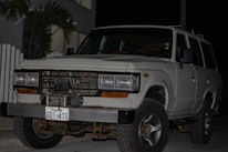 A white Toyota SUV is parked outdoors during the evening or night. The vehicle's front grille and headlights are clearly visible. It has a front-mounted license plate from Ecuador with the number GDI-419, and the front bumper appears robust. There are some palm leaves and a modern building in the background.