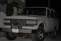 A white Toyota SUV is parked outdoors during the evening or night. The vehicle's front grille and headlights are clearly visible. It has a front-mounted license plate from Ecuador with the number GDI-419, and the front bumper appears robust. There are some palm leaves and a modern building in the background.