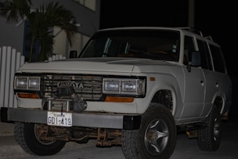 A white Toyota SUV is parked outdoors during the evening or night. The vehicle's front grille and headlights are clearly visible. It has a front-mounted license plate from Ecuador with the number GDI-419, and the front bumper appears robust. There are some palm leaves and a modern building in the background.