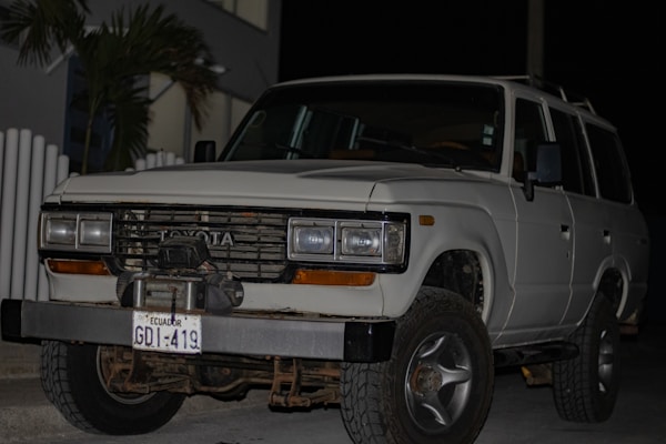 A white Toyota SUV is parked outdoors during the evening or night. The vehicle's front grille and headlights are clearly visible. It has a front-mounted license plate from Ecuador with the number GDI-419, and the front bumper appears robust. There are some palm leaves and a modern building in the background.
