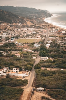 Aerial view of a coastal town with a mix of residential and undeveloped areas. The landscape features a winding road leading through the community, houses interspersed among greenery, and a backdrop of hills. The coastline, with waves gently lapping at the shore, stretches into the distance under a cloudy sky.