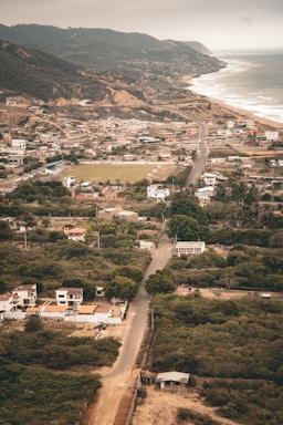 Aerial view of a coastal town with a mix of residential and undeveloped areas. The landscape features a winding road leading through the community, houses interspersed among greenery, and a backdrop of hills. The coastline, with waves gently lapping at the shore, stretches into the distance under a cloudy sky.