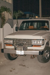 A white Toyota SUV with an Ecuadorian license plate is parked on a concrete surface at night. The vehicle appears to be an older model with noticeable wear on the bumper and front grille. There is a white picket fence and some palm trees in the background.