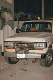 A white Toyota SUV with an Ecuadorian license plate is parked on a concrete surface at night. The vehicle appears to be an older model with noticeable wear on the bumper and front grille. There is a white picket fence and some palm trees in the background.