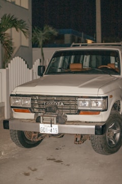 A white Toyota SUV with an Ecuadorian license plate is parked on a concrete surface at night. The vehicle appears to be an older model with noticeable wear on the bumper and front grille. There is a white picket fence and some palm trees in the background.