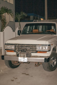 A white Toyota SUV with an Ecuadorian license plate is parked on a concrete surface at night. The vehicle appears to be an older model with noticeable wear on the bumper and front grille. There is a white picket fence and some palm trees in the background.