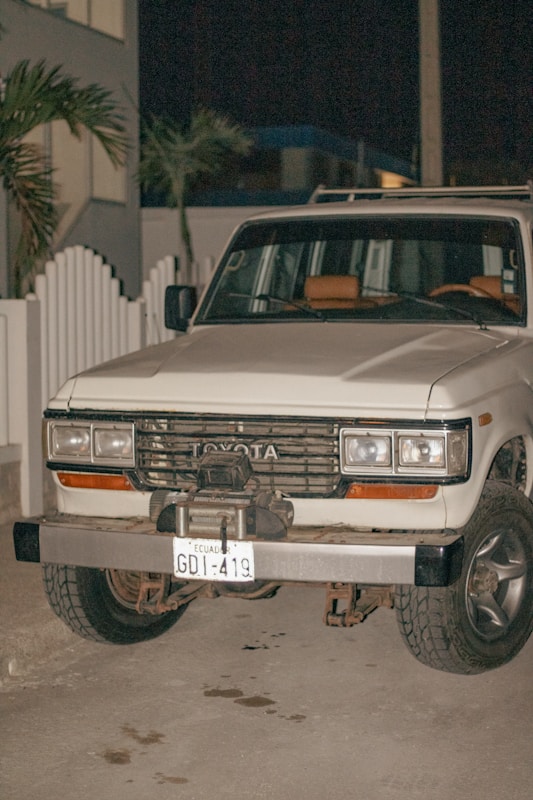A white Toyota SUV with an Ecuadorian license plate is parked on a concrete surface at night. The vehicle appears to be an older model with noticeable wear on the bumper and front grille. There is a white picket fence and some palm trees in the background.