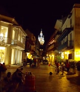 A cobblestone street is lined with colonial-style buildings illuminated by warm, yellow streetlights. People are walking, and some are gathered in groups, creating a lively but serene atmosphere. In the distance, a beautifully lit cathedral tower rises above the rooftops.