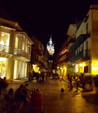 A cobblestone street is lined with colonial-style buildings illuminated by warm, yellow streetlights. People are walking, and some are gathered in groups, creating a lively but serene atmosphere. In the distance, a beautifully lit cathedral tower rises above the rooftops.