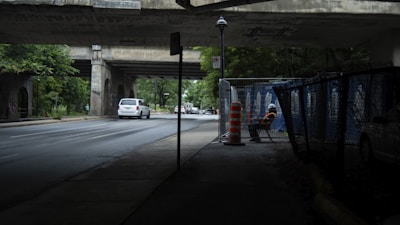 A road runs beneath a bridge, with a white vehicle driving on it. A worker in a reflective vest and hard hat is seated on a chair beside a construction fence, near an orange traffic barrel. There is graffiti visible on the bridge, and trees line the edges, with a small signpost in the middle of the scene.