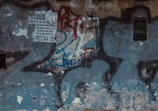 A dilapidated wall with several layers of peeling paint and graffiti. A printed advertisement, slightly torn, is partially visible in the upper left corner. The advertisement has text and a phone number. The wall appears weathered and neglected.