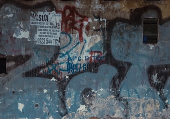 A dilapidated wall with several layers of peeling paint and graffiti. A printed advertisement, slightly torn, is partially visible in the upper left corner. The advertisement has text and a phone number. The wall appears weathered and neglected.