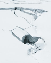 A landscape of a glacier featuring prominent cracks and crevices. The snowy surface appears rugged with jagged formations and deep gaps, suggesting a cold and harsh environment.