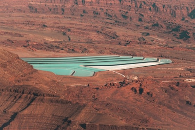 An aerial view of a series of turquoise evaporation ponds set in a reddish-brown desert landscape. The ponds are rectangular with visible white borders, contrasting sharply with the surrounding terrain. The landscape is characterized by rugged rock formations, creating a stark and barren feel.