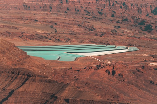 An aerial view of a series of turquoise evaporation ponds set in a reddish-brown desert landscape. The ponds are rectangular with visible white borders, contrasting sharply with the surrounding terrain. The landscape is characterized by rugged rock formations, creating a stark and barren feel.