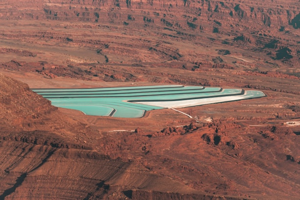 An aerial view of a series of turquoise evaporation ponds set in a reddish-brown desert landscape. The ponds are rectangular with visible white borders, contrasting sharply with the surrounding terrain. The landscape is characterized by rugged rock formations, creating a stark and barren feel.