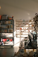 A warmly lit stationery shop interior features shelves lined with books and colorful bags, a display of greeting cards arranged on the wall, and various stationery items organized neatly. Sunlight casts soft shadows across the floor.
