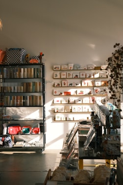 A warmly lit stationery shop interior features shelves lined with books and colorful bags, a display of greeting cards arranged on the wall, and various stationery items organized neatly. Sunlight casts soft shadows across the floor.