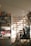 A warmly lit stationery shop interior features shelves lined with books and colorful bags, a display of greeting cards arranged on the wall, and various stationery items organized neatly. Sunlight casts soft shadows across the floor.