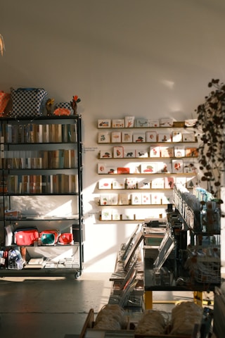 A warmly lit stationery shop interior features shelves lined with books and colorful bags, a display of greeting cards arranged on the wall, and various stationery items organized neatly. Sunlight casts soft shadows across the floor.