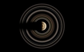 A celestial object with prominent, concentric rings surrounding a central, round figure. The rings display varying shades and thicknesses, with the central sphere showing a textured surface.