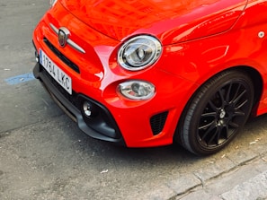 A close-up of the front section of a vibrant red compact car, showcasing its sleek design, prominent headlight, and glossy paint. The car features a detailed emblem and a license plate attached to the front.