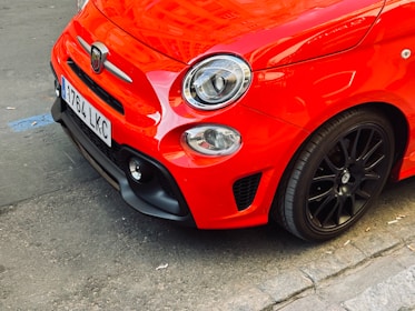 A close-up of the front section of a vibrant red compact car, showcasing its sleek design, prominent headlight, and glossy paint. The car features a detailed emblem and a license plate attached to the front.