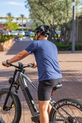 A person wearing a blue shirt and black shorts is sitting on an electric mountain bike on a paved walkway. The cyclist is equipped with a black helmet, and the bike is branded Kingbull. In the background, there are trees, parked cars, and buildings, which suggest an urban environment with greenery.