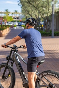A person wearing a blue shirt and black shorts is sitting on an electric mountain bike on a paved walkway. The cyclist is equipped with a black helmet, and the bike is branded Kingbull. In the background, there are trees, parked cars, and buildings, which suggest an urban environment with greenery.