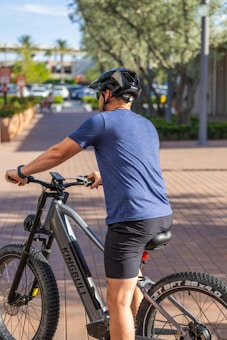 A person wearing a blue shirt and black shorts is sitting on an electric mountain bike on a paved walkway. The cyclist is equipped with a black helmet, and the bike is branded Kingbull. In the background, there are trees, parked cars, and buildings, which suggest an urban environment with greenery.