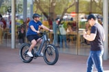A student weaving through campus pathways on a sleek black bike with electric blue highlights, city buildings blurred behind.