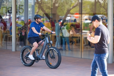 A student weaving through campus pathways on a sleek black bike with electric blue highlights, city buildings blurred behind.