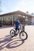 A man wearing a blue t-shirt and shorts stands with a bicycle on a brick-paved area in front of a large glass-fronted store. The store has a modern design with clean lines and a prominent logo. The man is wearing a helmet and sunglasses, and appears relaxed. People are visible inside the store and in the background outside.