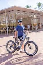 A person is standing outside a modern glass-fronted store, holding a large mountain bike. The person is wearing a blue shirt, black shorts, a helmet, and sunglasses. The bike is a silver color with thick tires. The store has a transparent facade and an Apple logo is visible, suggesting it's an Apple Store. Palm trees are in the background, indicating a warm climate.