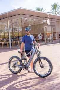 A person is standing outside a modern glass-fronted store, holding a large mountain bike. The person is wearing a blue shirt, black shorts, a helmet, and sunglasses. The bike is a silver color with thick tires. The store has a transparent facade and an Apple logo is visible, suggesting it's an Apple Store. Palm trees are in the background, indicating a warm climate.