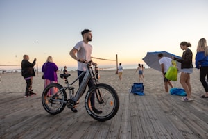 A group of people are enjoying a beach setting during sunset. A man stands next to an electric bicycle on a wooden boardwalk. Others are either walking or standing with various beach accessories like bags and an umbrella. Seagulls are flying in the sky, and a volleyball game is happening in the background.