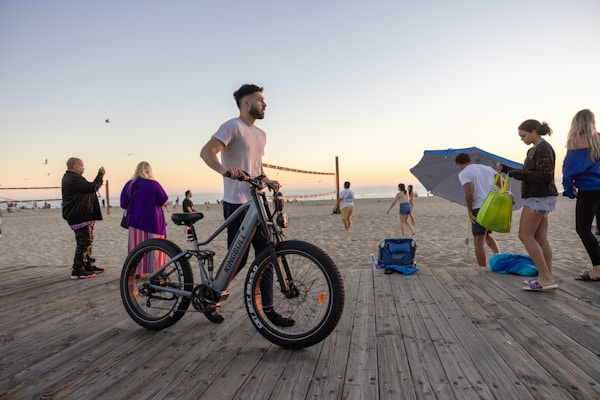 A group of people are enjoying a beach setting during sunset. A man stands next to an electric bicycle on a wooden boardwalk. Others are either walking or standing with various beach accessories like bags and an umbrella. Seagulls are flying in the sky, and a volleyball game is happening in the background.