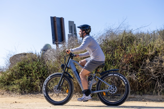 A person wearing a helmet and casual clothing is riding an electric bike on a dirt path. The bike features large, sturdy tires and the brand name 'Kingbull' visible on the frame. The background includes dry vegetation and a clear blue sky.