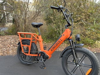 An orange electric bike with the brand name 'KINGBULL' is parked on a paved path. The bike features large, thick tires and a sturdy frame. Surrounding the bike are trees and bushes with autumn leaves scattered on the ground, creating a natural backdrop.