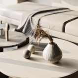 A minimalist interior scene featuring a beige sofa with a checkered grey and white blanket draped over it. In the foreground, there are two light wooden coffee tables. One table has a white vase with dried grass, and the other has two spherical decorative objects and a cup.