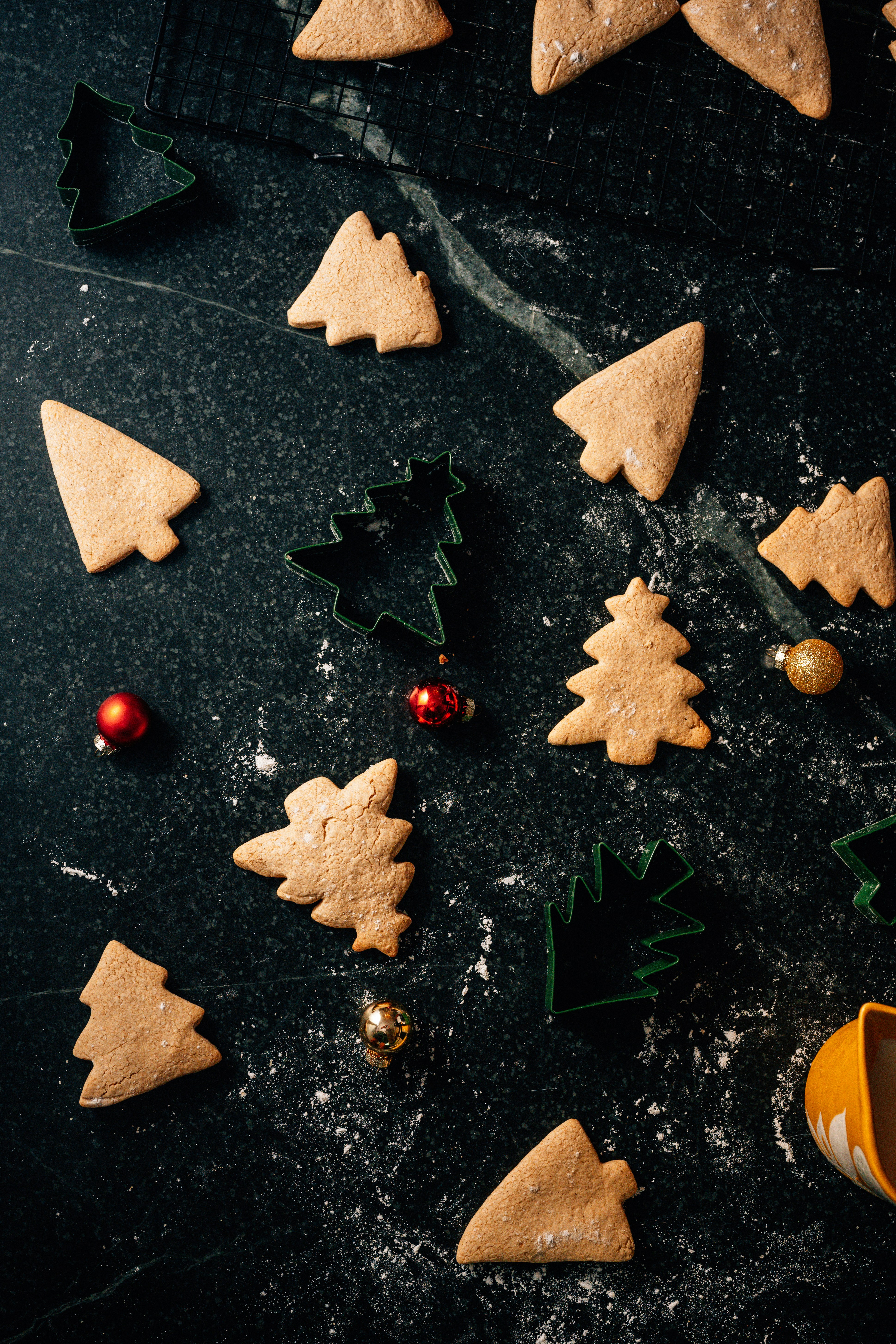 Several tree-shaped cookies and cookie cutters arranged on a dark marble surface. Small, colorful Christmas ornaments are scattered among the cookies. The cookies are light brown and dusted with a bit of flour, providing a festive baking scene.