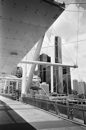 An urban scene featuring a modern suspension bridge with geometric structures and cables, leading towards a backdrop of towering skyscrapers. The architectural elements of the bridge create an intricate pattern, while the high-rise buildings stand prominently in the background.